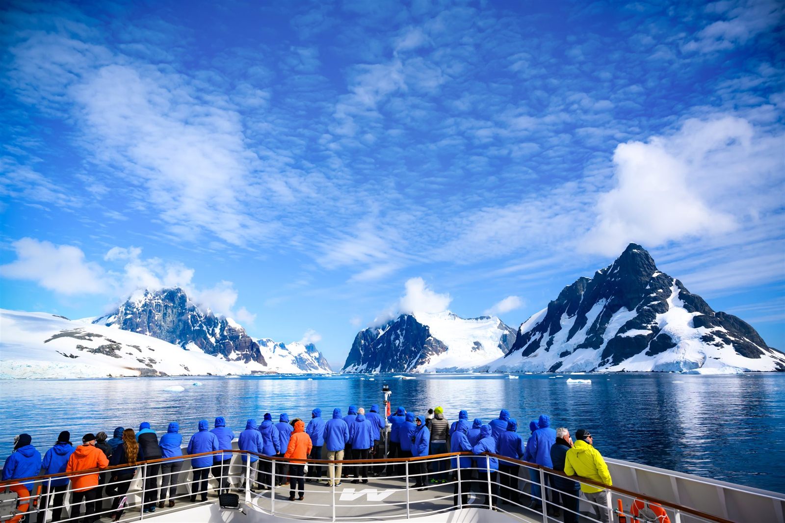 Passengers in parkas on the bow of a small ship facing snowy polar peaks